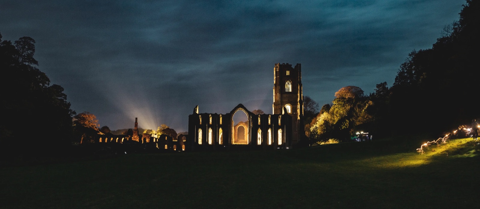 Fountains Abbey after dark, its tower and arched windows lit gold from inside against a deep blue night sky.