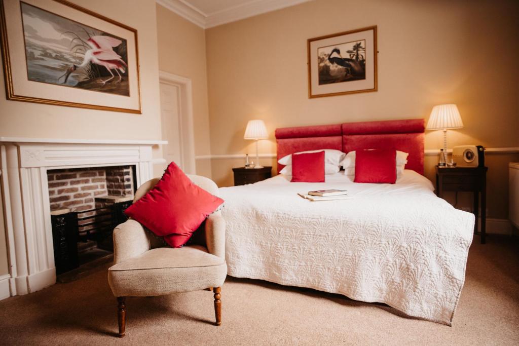 A bedroom with a red upholstered headboard, a white fireplace, Audubon bird prints and a small armchair.