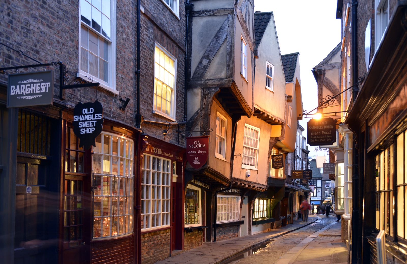 The Shambles in York at dusk: leaning timber-framed shopfronts with lit windows along a narrow stone street.
